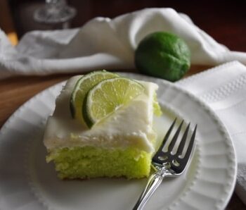 A square piece of light green key lime cake with cream cheese icing with 2 half lime slices on top; served on a scalloped round white plate. On the plate is a sparkling dinner fork. Beside it is a textured cloth white napkin, and in the background is one fresh lime.