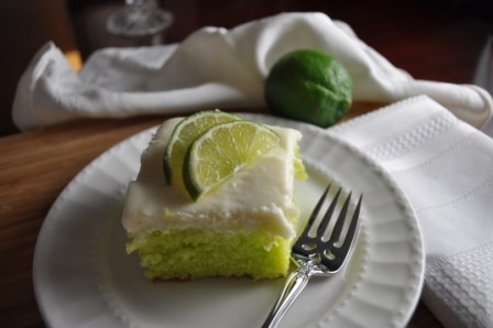 A square piece of light green key lime cake with cream cheese icing with 2 half lime slices on top; served on a scalloped round white plate. On the plate is a sparkling dinner fork. Beside it is a textured cloth white napkin, and in the background is one fresh lime.