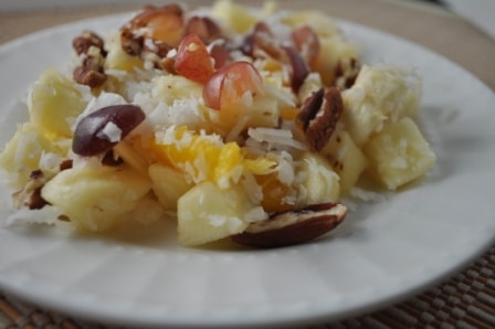 Fruit salad with peeled apples, bananas, red grapes, orange slices, coconut and pecans on a white dessert plate on a straw placemat.