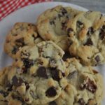 Six chocolate chip cookies with nuts piled on a round white plate with a red and white checked napkin in top left.