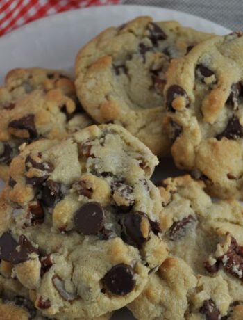 Six chocolate chip cookies with nuts piled on a round white plate with a red and white checked napkin in top left.
