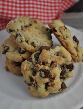 Chocolate Chip cookies stacked and balanced on a white plate with a red and white checkered tablecloth in top left corner.