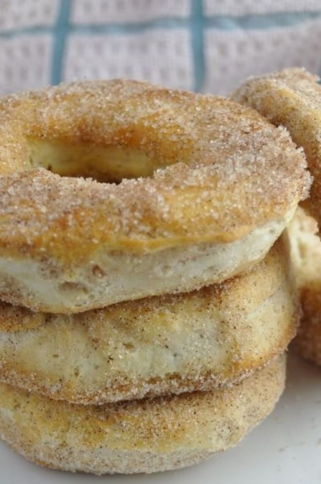 Close up picture of three delicious looking donuts made from canned biscuits in the air fryer and coated with crystal white sugar mixed with brown cinnamon. Arranged donuts are sitting on a plate stacked three high with one additional donut on the right side of the picture leaning leftwards on the stack of donuts. White cloth with blue lines forming squares is blurred in the background at the top of the picture.