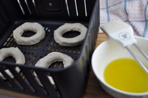 Four canned grand biscuits with the centers cut out to leave a hole for donuts that are sitting in a black air fryer basket. Olive oil in a small bowl with a brush on top on right side of picture.
