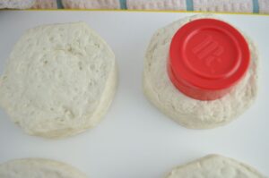 Biscuit dough on a cutting board with top right biscuit having a spice bottle top in the middle to cut out a hole for donuts.