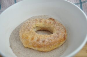 One hot doughnut made from biscuit dough in the air fryer sitting in a white bowl that contains cinnamon and sugar mixture to coat the donut.