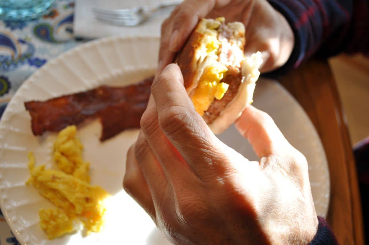 Hands holding a sausage egg biscuit that was part of Blackstone Breakfast Recipes prepared on the Blackstone griddle with scrambled eggs and bacon on a serving plate.