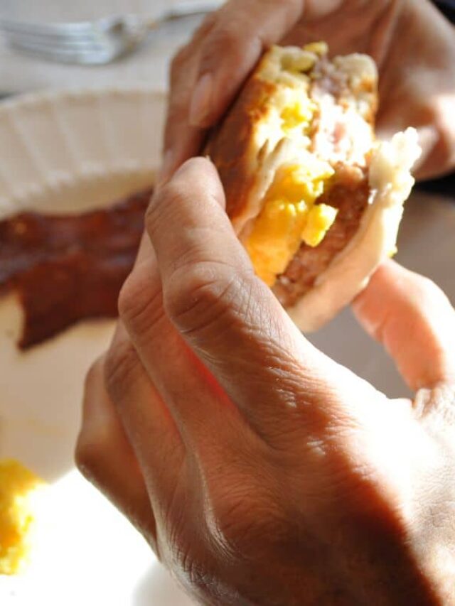 Hands holding a sausage egg biscuit that was part of Blackstone Breakfast Recipes prepared on the Blackstone griddle with scrambled eggs and bacon on a serving plate.