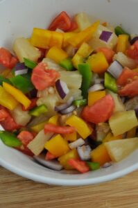 A mixture of golden pineapple chunks, chopped red and green bell peppers, chopped red onions with a deep purple outside and crisp white inside, fresh tomato tiny cubes all in a white bowl.